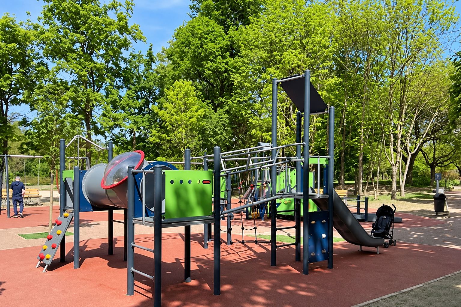 Une structure de jeux en métal bleu foncé et vert avec toboggan et tunnel, installée sur un sol souple rouge dans un parc ombragé.