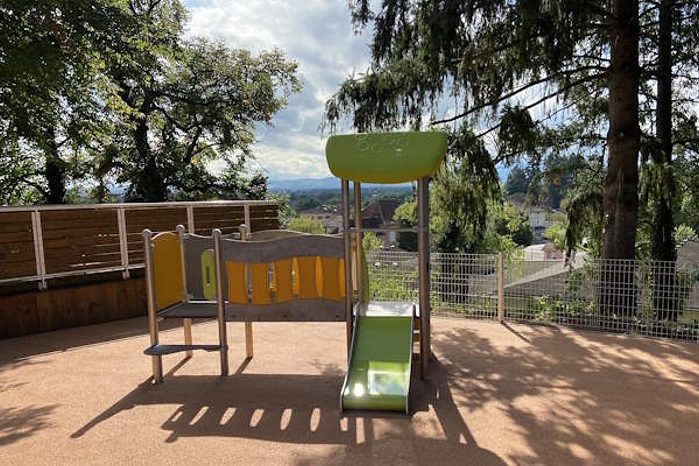 Une petite structure de jeux avec toboggan vert posée sur un sol souple beige, entourée de grands arbres, avec une vue dégagée sur la campagne.