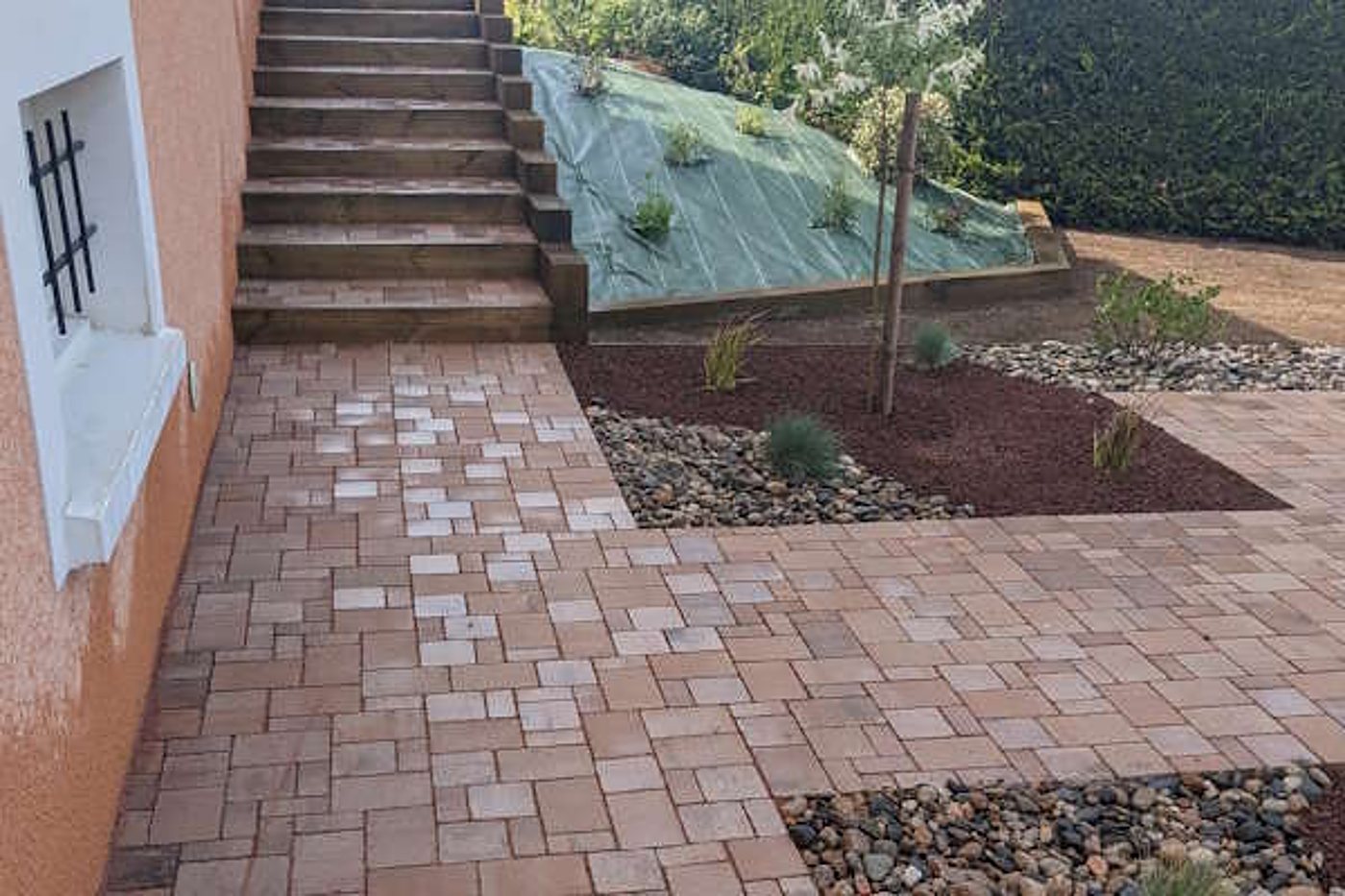 Une allée en pavés rosés longe un escalier en bois et des massifs de paillage rouge et de galets plantés de jeunes végétaux.
