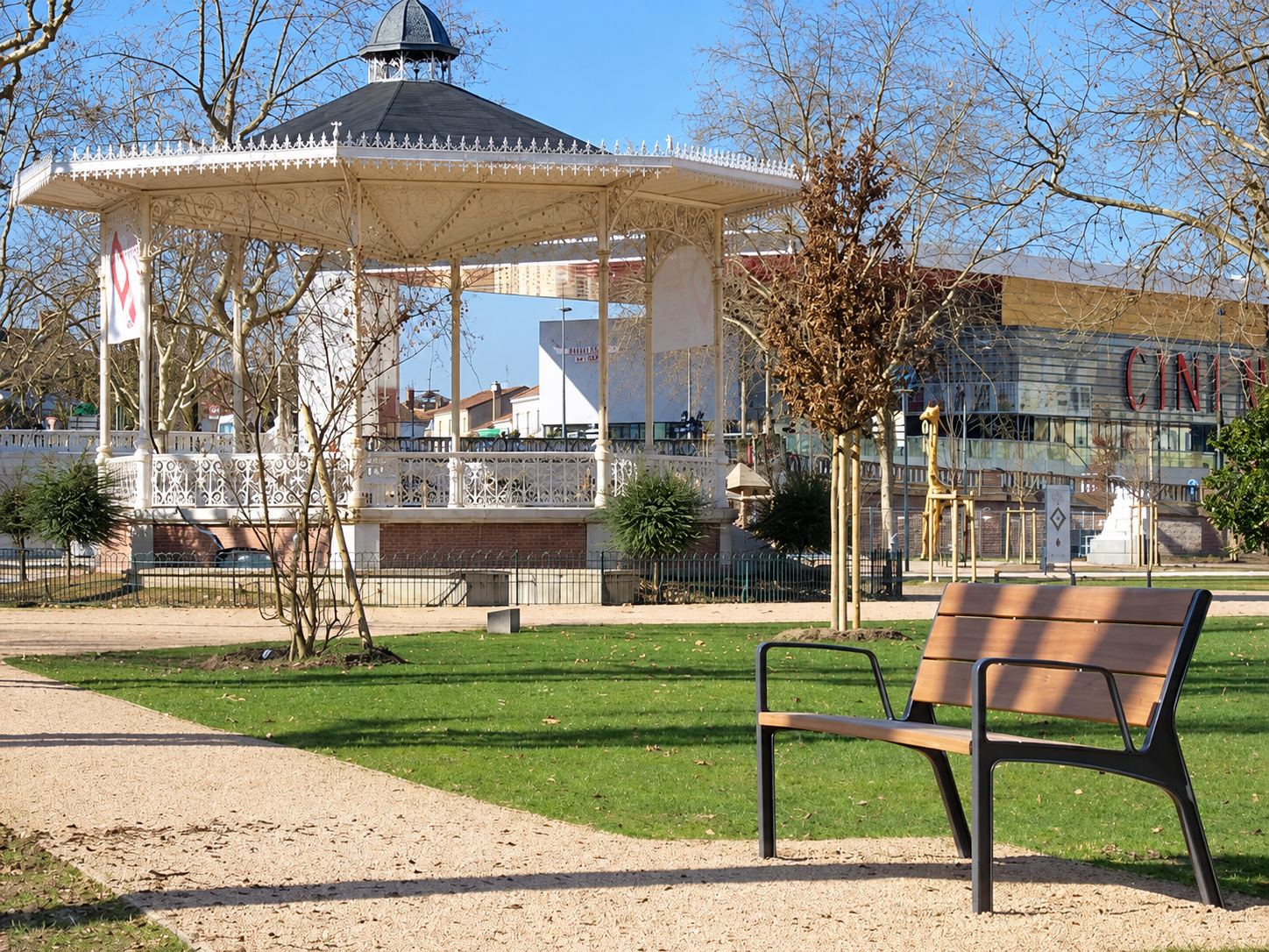 Un banc moderne en bois et métal posé sur une pelouse, face à un kiosque à musique ancien et à un cinéma dans un parc public ensoleillé.