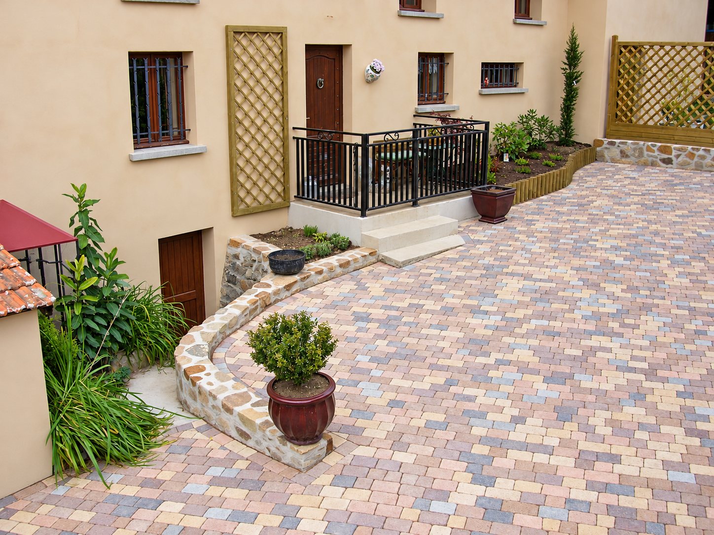 Une cour pavée de pavés multicolores aux tons beige, rose et gris, bordée d'un muret en pierre, de pots de plantes et d'un escalier menant à une terrasse avec garde-corps devant une façade crépie.