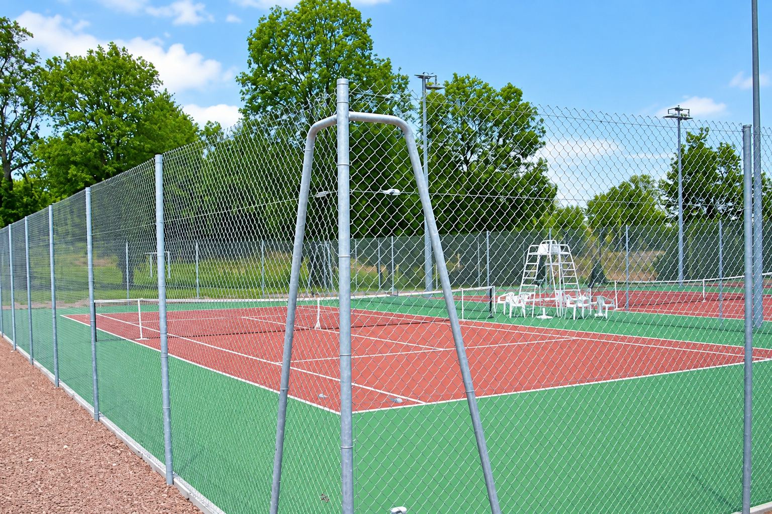 Un court de tennis extérieur rouge et vert, entouré d'un haut grillage métallique, avec une chaise d'arbitre et des chaises blanches au fond.