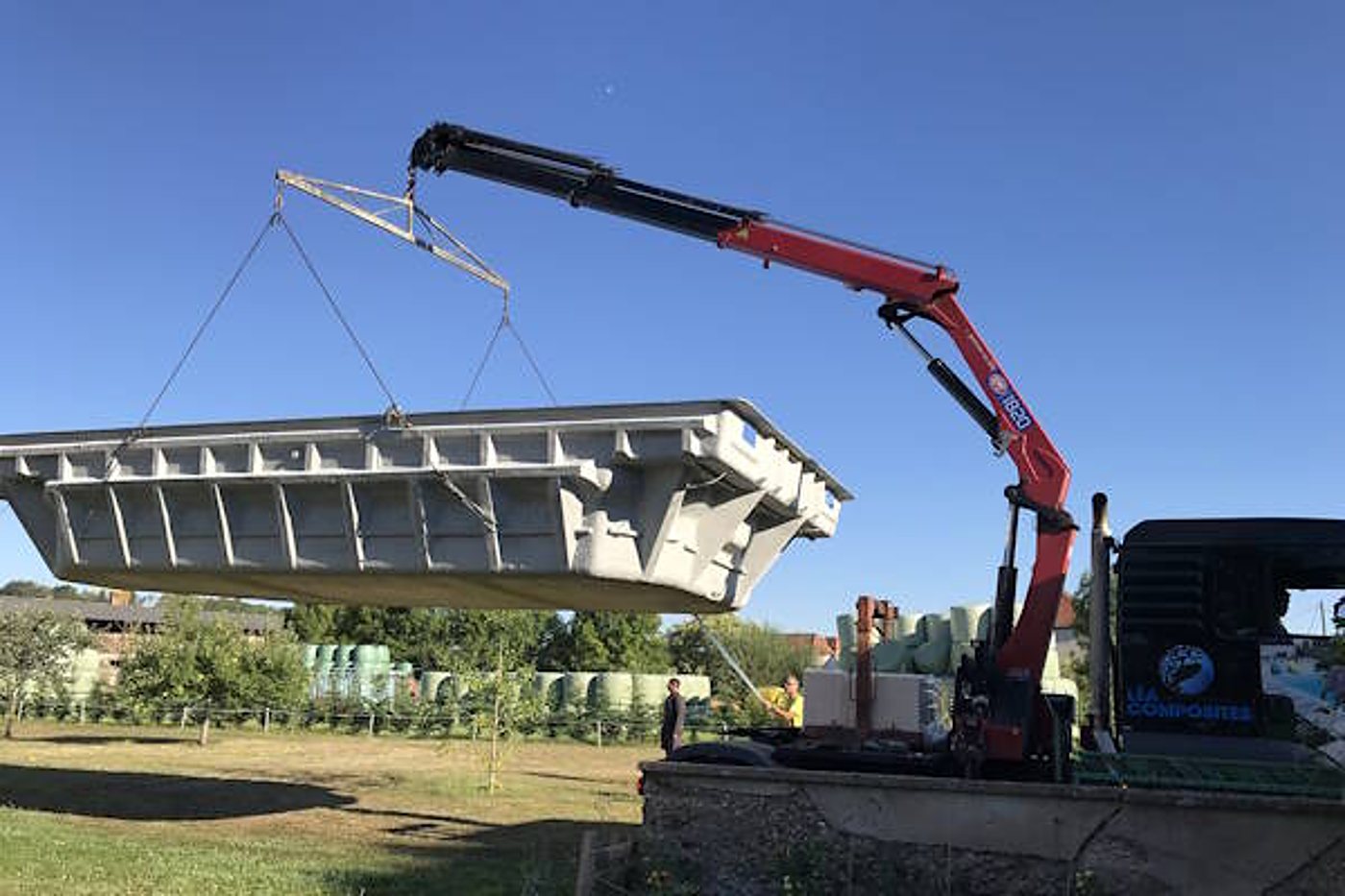 Un camion équipé d'une grue rouge soulève une grande coque de piscine grise au-dessus d'un terrain, sous un ciel bleu.