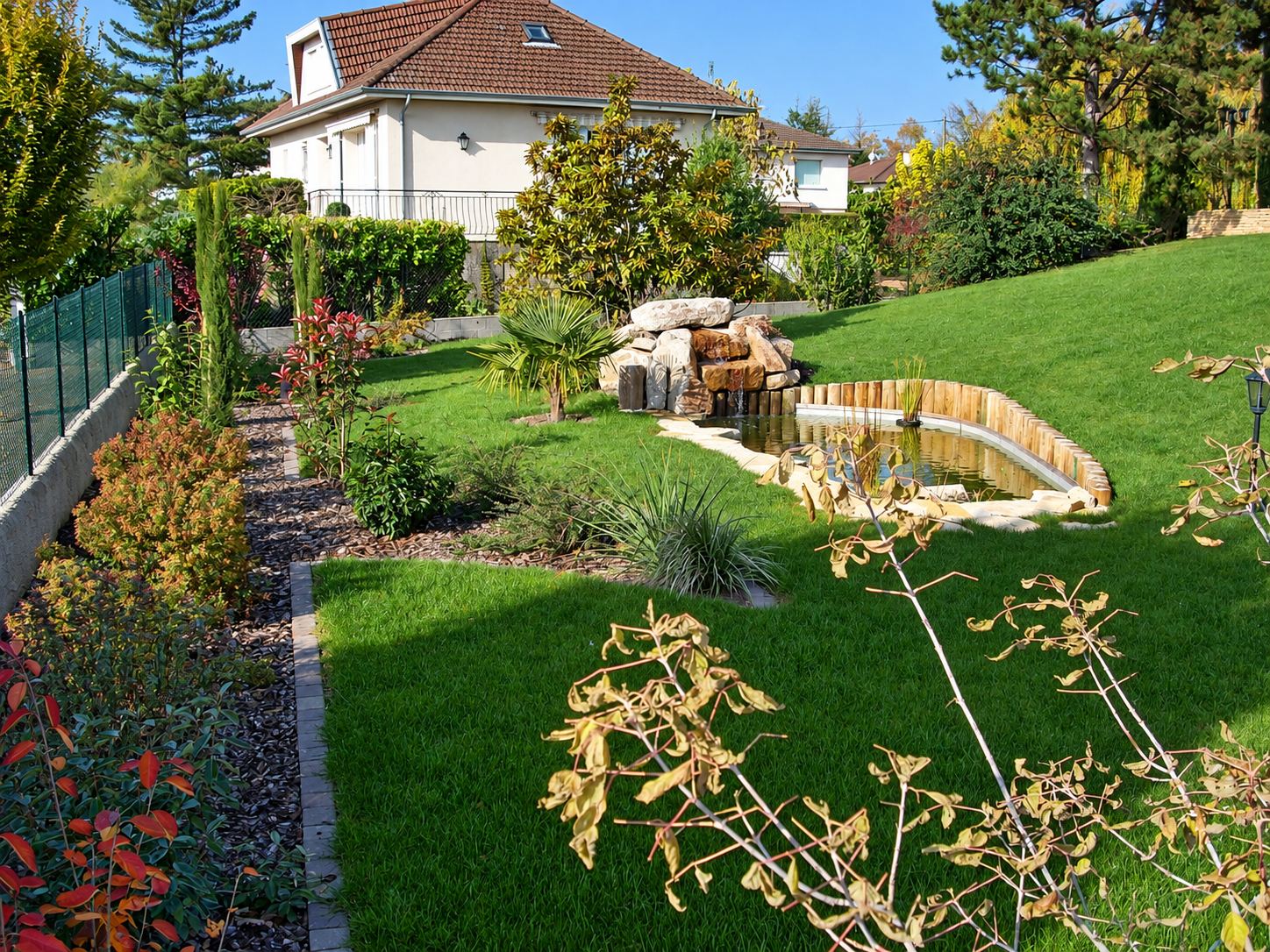 Jardin aménagé en pente avec pelouse verte, massifs d'arbustes, bassin bordé de rondins de bois et cascade de pierres devant une maison blanche.