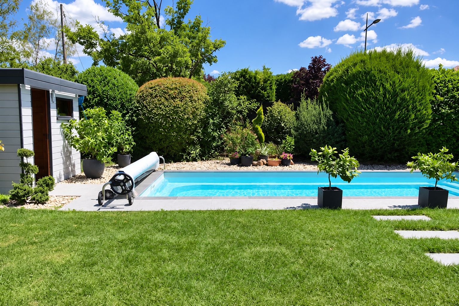 Une piscine rectangulaire à l'eau turquoise avec son enrouleur de bâche, entourée d'une pelouse, de haies taillées, de plantes en pots et d'un abri de jardin gris.