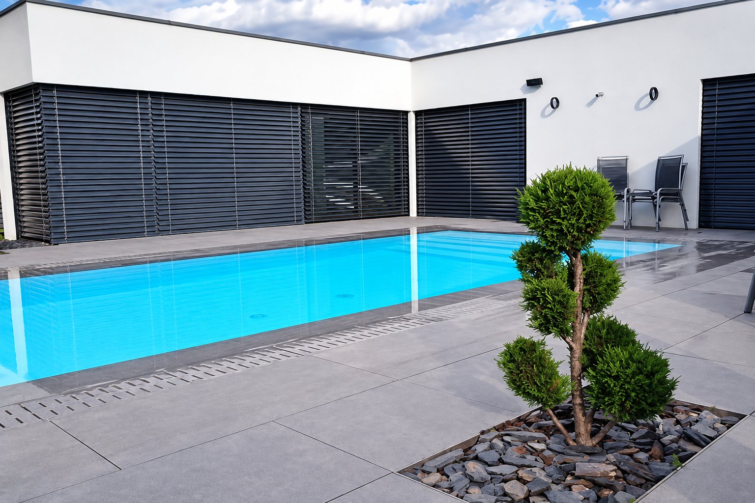 Une piscine rectangulaire à l'eau turquoise bordée de larges dalles grises, devant une maison blanche à volets noirs, avec un thuya taillé en nuage au premier plan.