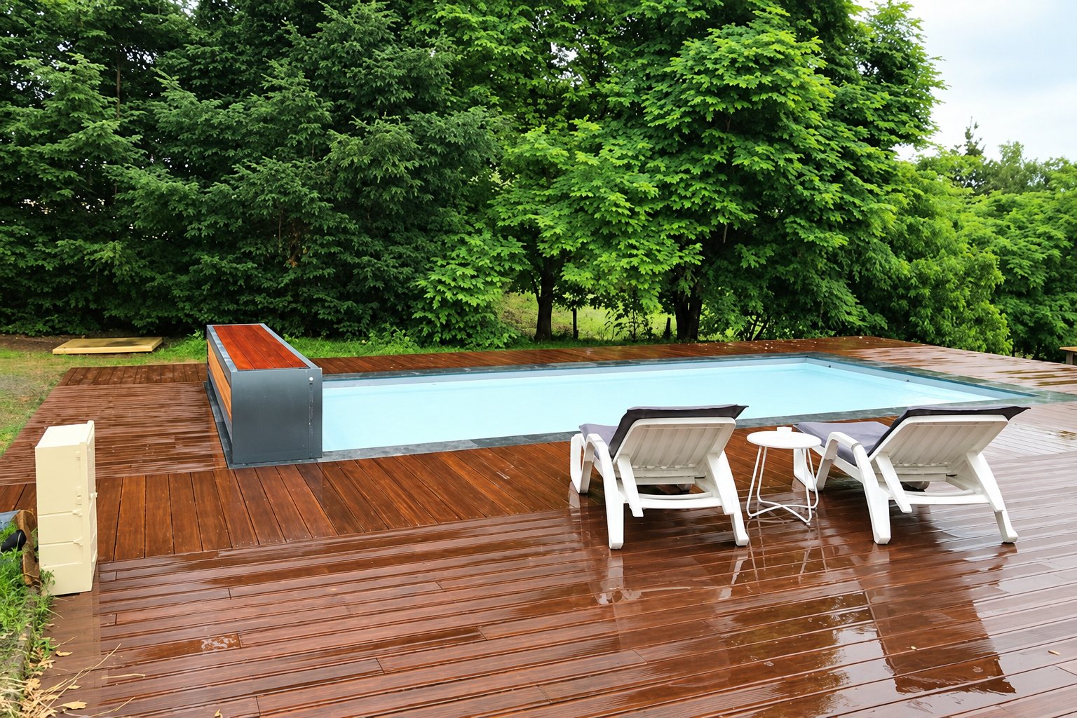 Une piscine rectangulaire à volet couvert entourée d'une terrasse en bois humide avec deux chaises longues blanches devant des arbres verts.