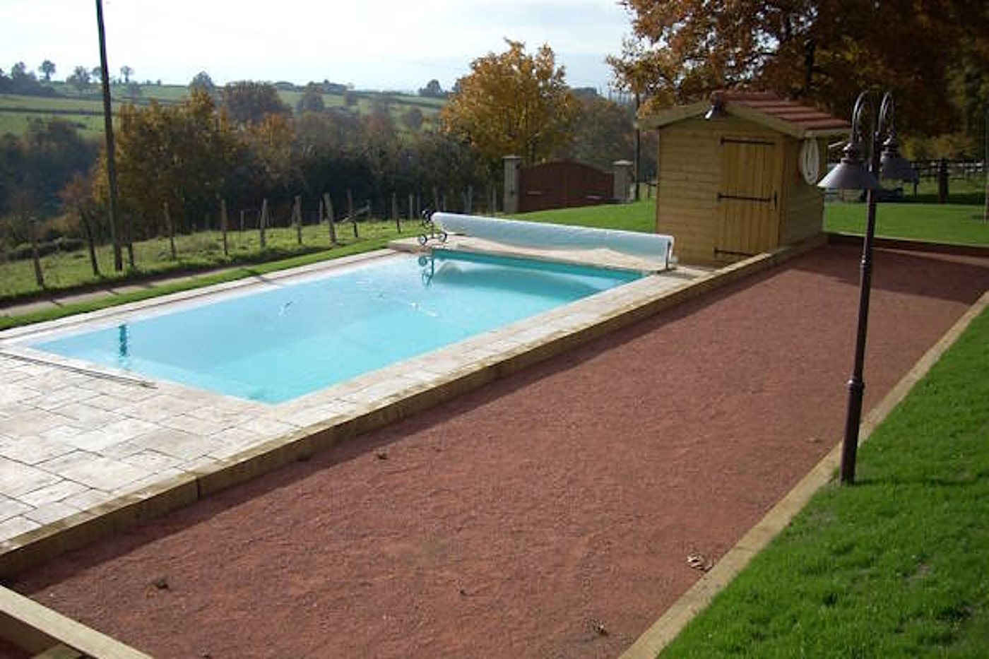 Une piscine rectangulaire à l'eau turquoise, bordée de dalles en pierre et d'une allée de gravier rouge, avec un abri de jardin en bois et un lampadaire dans un jardin de campagne.