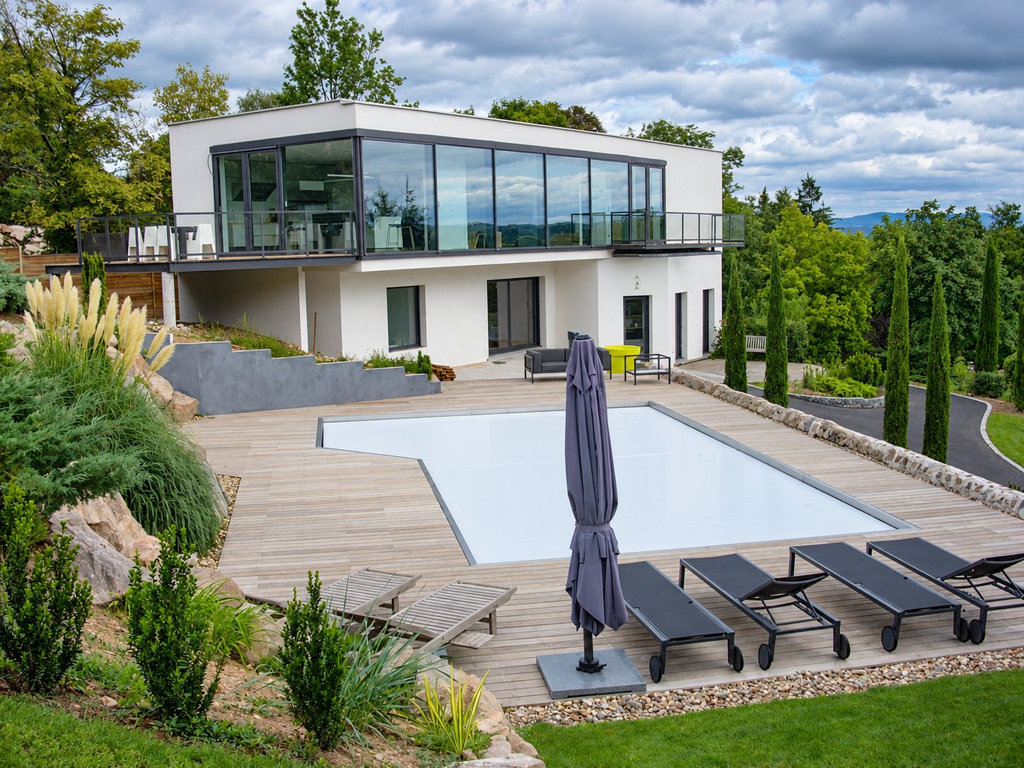 Une piscine fermée par un volet blanc, entourée d'une terrasse en bois avec des bains de soleil et un parasol, devant une maison contemporaine à étage.