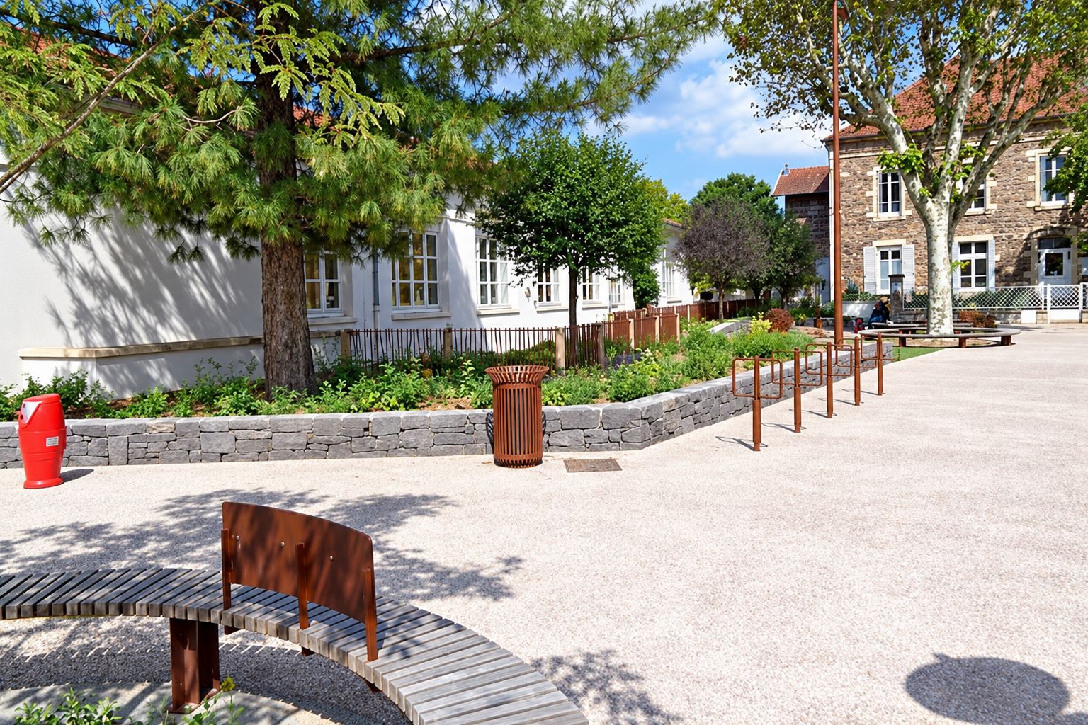 Place publique récemment aménagée avec un banc courbe en bois, un muret en pierre bordant des massifs de plantes, une poubelle et des arbres, devant des bâtiments en pierre.