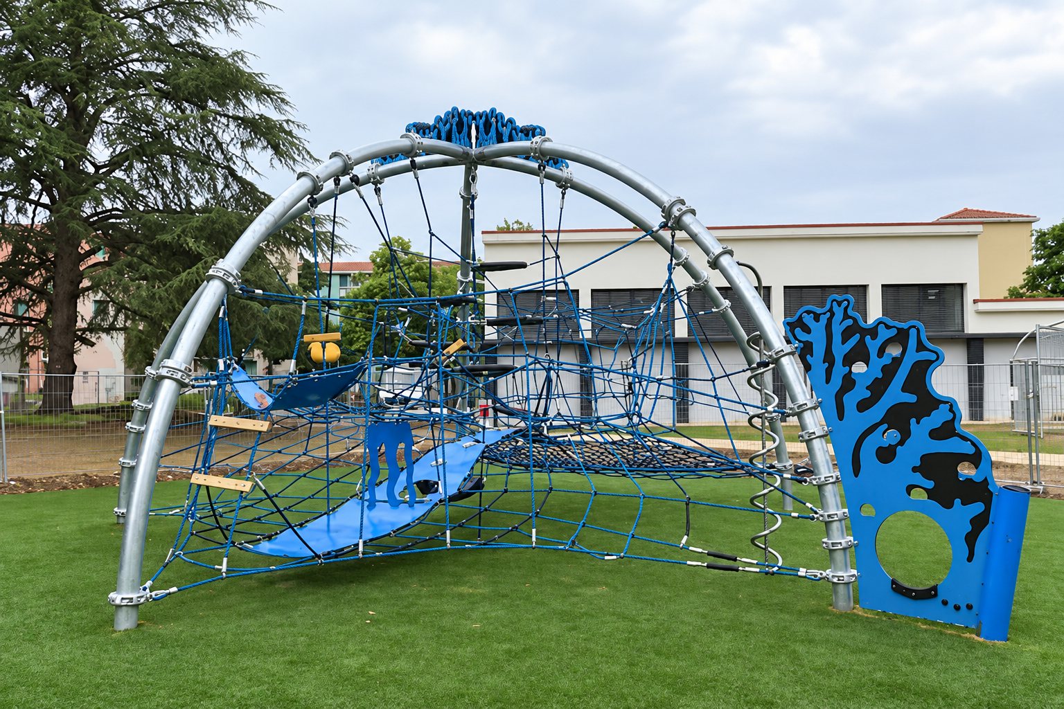 Une grande structure de jeux pour enfants avec arceaux en métal, filets bleus et toboggan bleu, installée sur une pelouse devant un bâtiment.