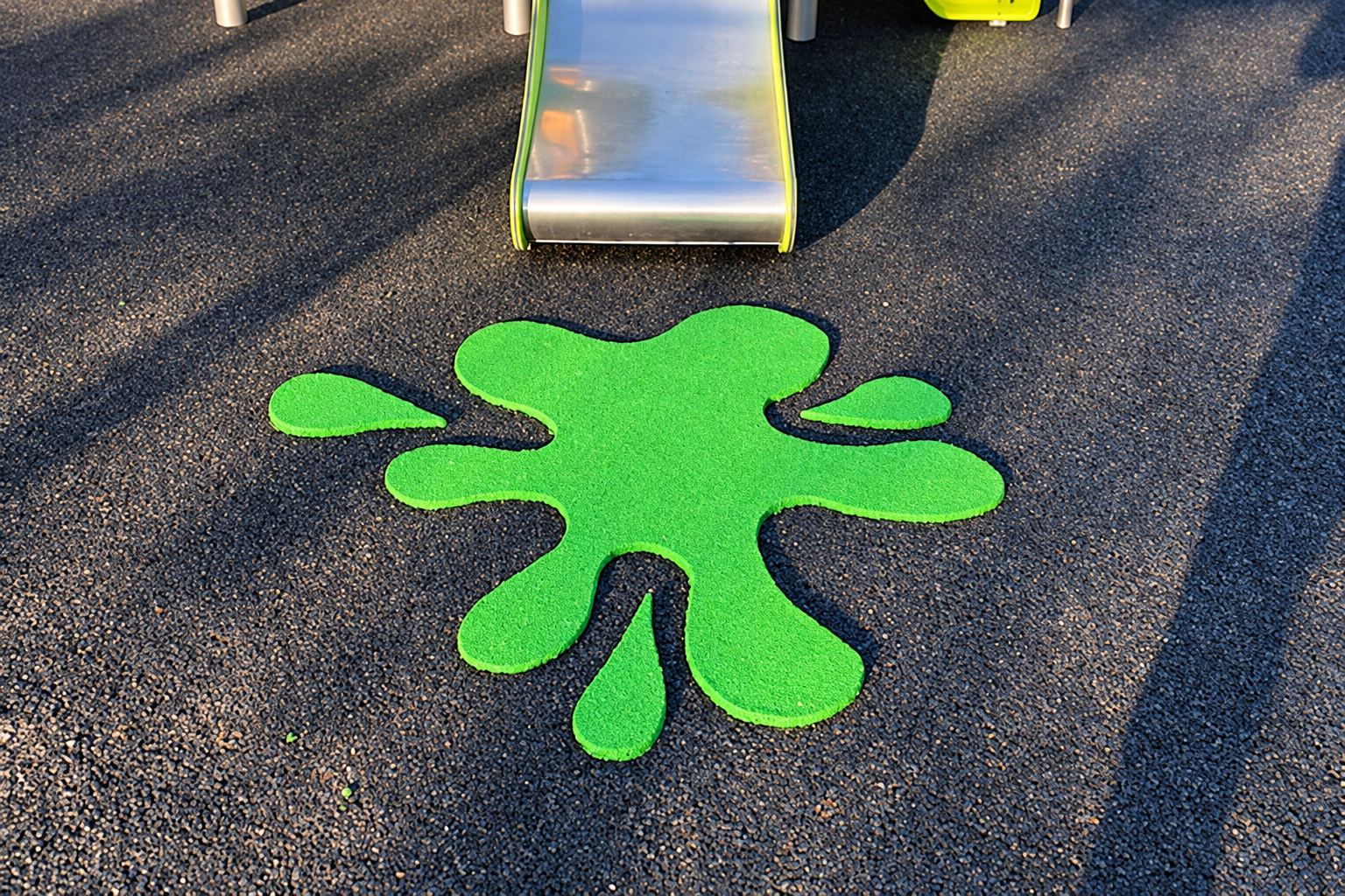 Une grande tache verte en forme d'éclaboussure est incrustée dans un revêtement de sol noir, devant un toboggan en métal sur une aire de jeux.