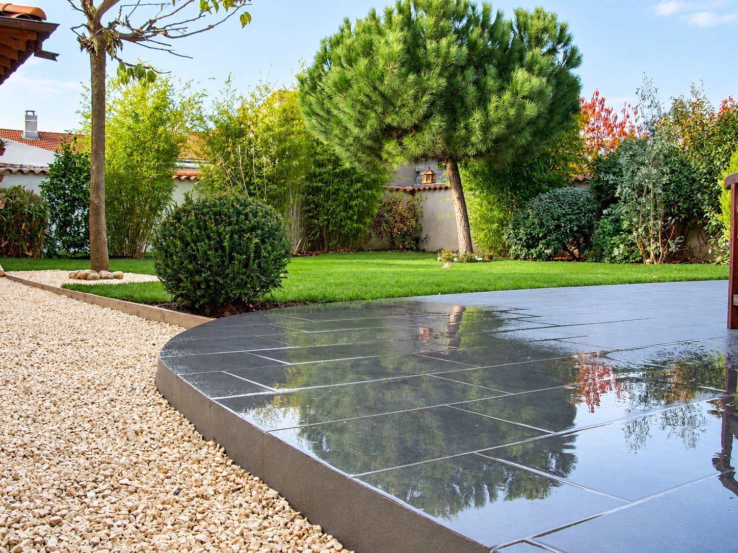 Une terrasse en dalles grises mouillées aux bords arrondis borde un jardin avec gravier clair, pelouse verte, arbustes et un pin parasol.