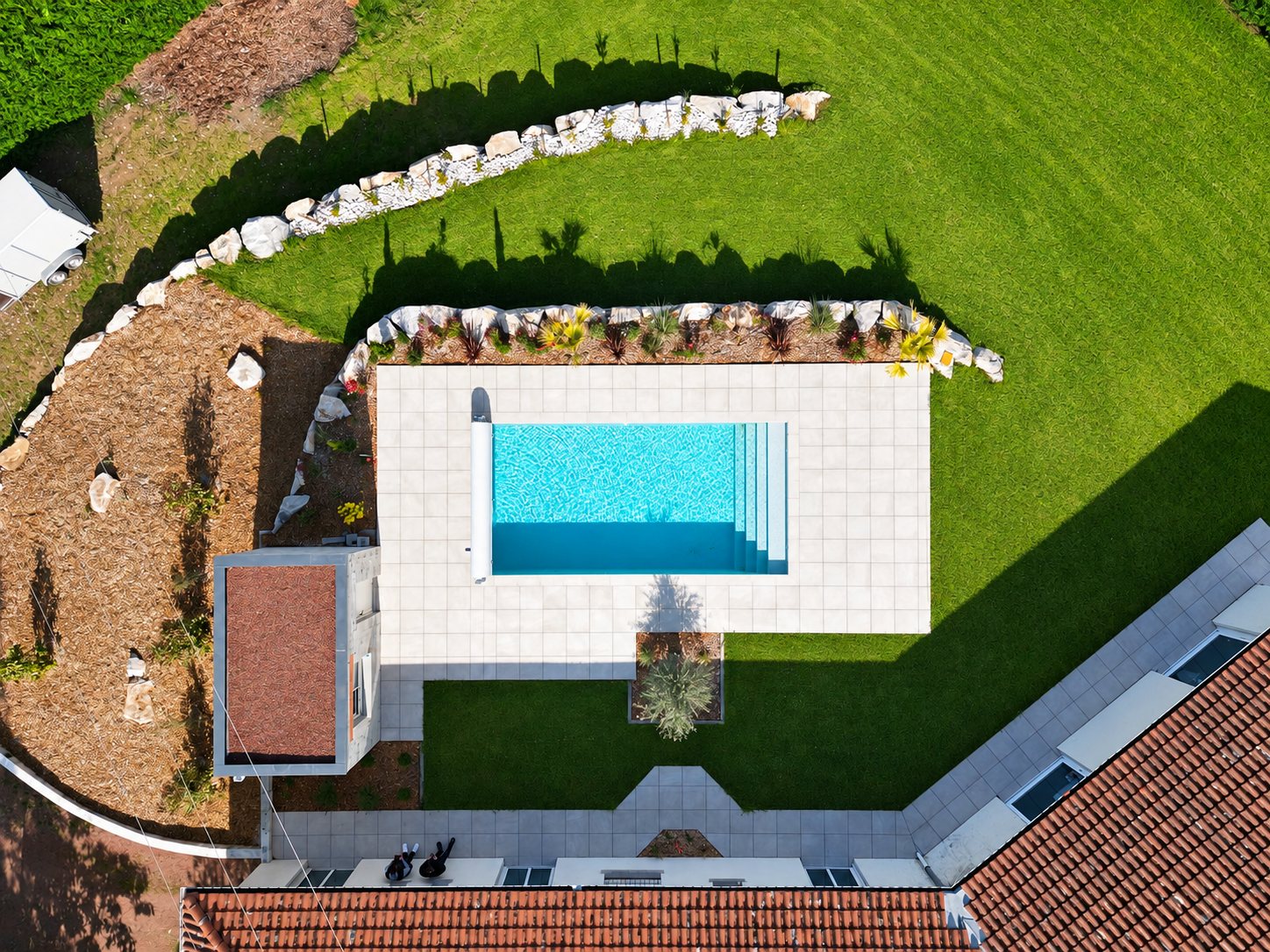 Vue de drone d'une piscine rectangulaire à l'eau turquoise, entourée d'une terrasse en dalles claires, de rochers et d'une pelouse.